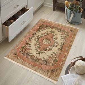 May include: A rectangular rug with a distressed, vintage look. The rug features a central cream-colored medallion surrounded by a floral pattern in shades of pink, green, and brown. The rug has a fringed edge and is placed on a light wood floor.