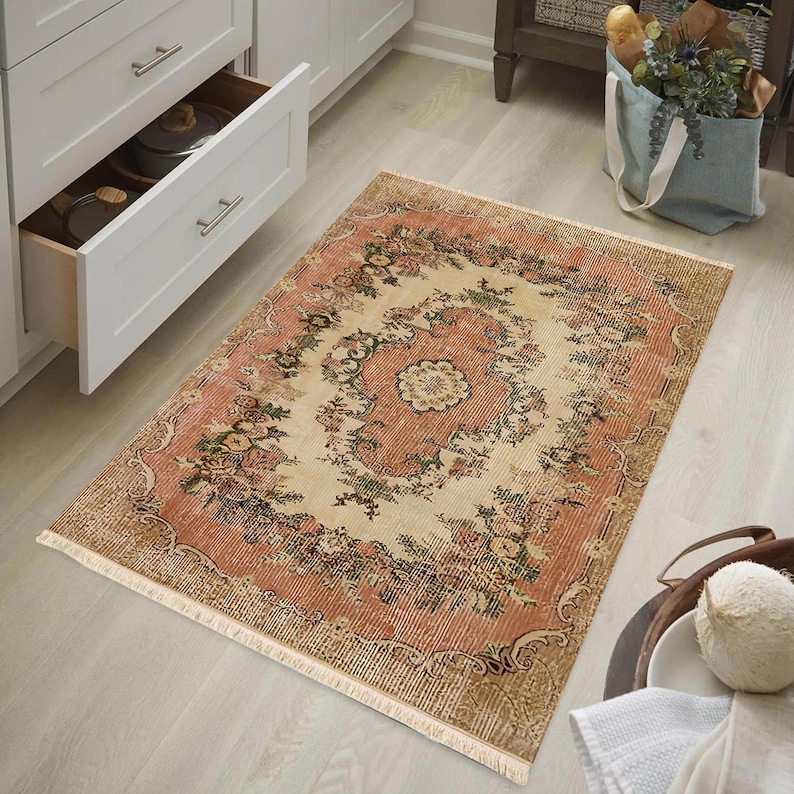 May include: A rectangular rug with a distressed, vintage look. The rug features a central cream-colored medallion surrounded by a floral pattern in shades of pink, green, and brown. The rug has a fringed edge and is placed on a light wood floor.