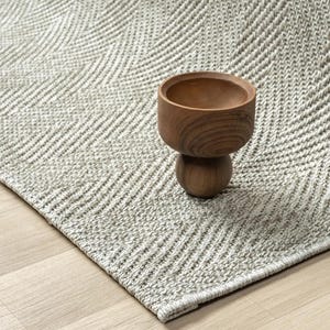 May include: A light grey rug with a woven chevron pattern. A small, wooden bowl with a rounded base sits on the rug. The bowl is made of a light brown wood and has a smooth finish. The rug is on a light wood floor.