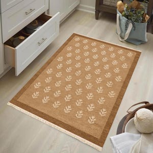 a rug with a leaf pattern on it, placed on a wooden floor in a kitchen setting. There are white cabinets and drawers in the background, as well as a blue bag and a bowl on the floor.