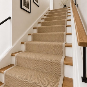 Op de afbeelding: Een trap met een beige geweven loper met een visgraatpatroon en lichtgekleurde stoffen randen. De loper bedekt het midden van de houten treden, geflankeerd door witte stootborden en een houten leuning met zwarte metalen steunen.