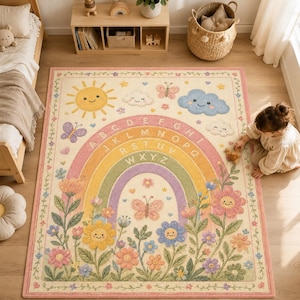 May include: A rectangular area rug featuring a cream background with a central rainbow arch containing the alphabet letters A through Z. The design includes smiling sun, cloud, and flower motifs, along with butterflies in pastel shades of pink, yellow, blue, and green, all framed by a decorative floral border.