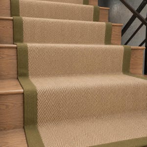 May include: A stair runner featuring a tan herringbone pattern with solid olive green fabric borders installed on wooden stairs. The runner covers the centre of each step and the riser, creating a continuous textured path up the staircase.