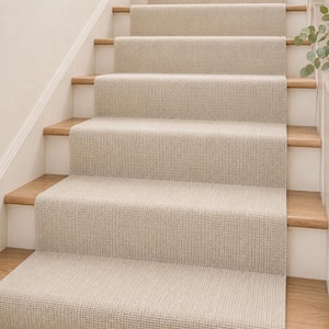May include: A staircase featuring light beige, textured carpet runner installed over natural wood treads. The carpet has a tight, woven grid pattern that covers the center of each step, contrasting with the white risers and light wood stair edges.