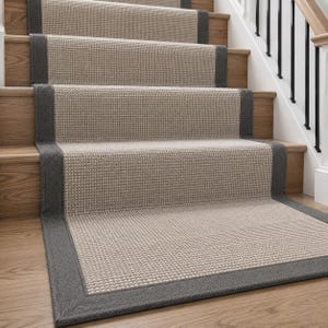 May include: A stair runner featuring a textured, light beige woven centre panel with a tight grid pattern. The runner is bordered by a wide, solid dark charcoal grey fabric trim that frames each step and the bottom landing of the wooden staircase.