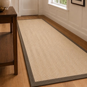 May include: A rectangular area rug featuring a light beige woven centre with a distinct herringbone pattern. The rug is framed by a wide, solid dark grey fabric border that runs along all four edges, creating a clean and defined rectangular shape.