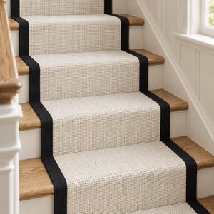 May include: A stair runner featuring a textured, light beige woven material with a solid black border running along both sides. The runner is installed over wooden stair treads, covering the centre portion of each step in a straight vertical line.