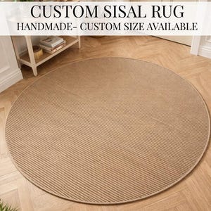 Puede incluir: Una alfombra redonda de sisal beige con una superficie texturizada y acanalada. La alfombra es hecha a mano y está disponible en tamaños personalizados. El texto "CUSTOM SISAL RUG HANDMADE- CUSTOM SIZE AVAILABLE" se muestra en la parte superior de la imagen.
