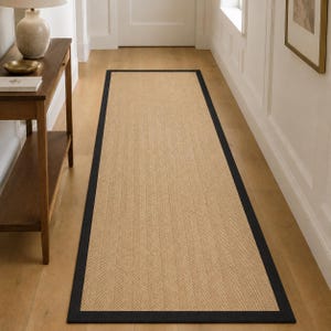 May include: A long rectangular runner rug featuring a natural tan woven texture with a subtle herringbone pattern. The rug is framed by a solid black border that runs along all four edges, creating a distinct contrast against the tan centre.