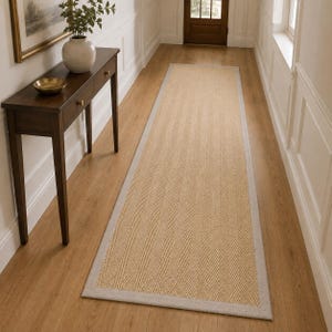 Op de afbeelding: Een lange, rechthoekige loper met een beige, geweven visgraatpatroon en een effen lichtgrijze rand. Het kleed ligt in de lengte op een houten vloer en strekt zich uit van de voorgrond naar de achterkant van de gangruimte.