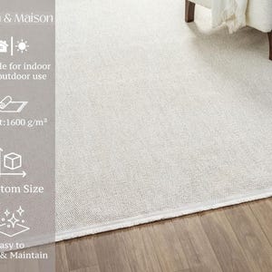 a white area rug on a wooden floor, with a chair visible in the background. The rug is described as suitable for indoor and outdoor use, with a weight of 1600 g/m². It also features a custom size and is easy to clean and maintain.