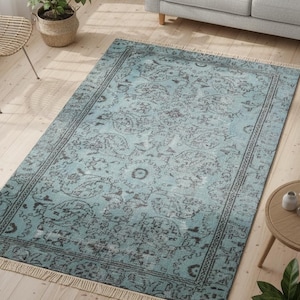 May include: A rectangular, turquoise area rug with a distressed, ornate pattern. The rug features a dark gray border and fringe along the short ends. The rug is placed on a light wood floor, with a sofa and side table in the background.