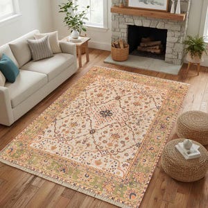 May include: An area rug with a cream-colored field and an intricate floral pattern in shades of orange, blue, and green. The rug has a green border and fringe along the edges. The rug is placed on a hardwood floor.