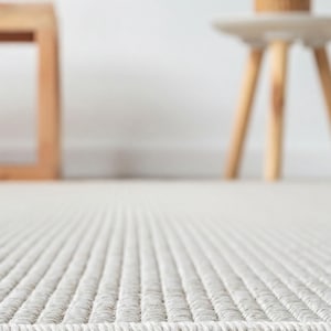 a white woven rug on a wooden floor, with a small wooden stool and a plant in a basket in the background.