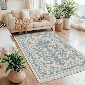 May include: A rectangular rug featuring a cream-coloured base with a light blue floral and scroll pattern. The rug has a border and fringing. It is placed on a wooden floor in a living room, complementing the room's decor.