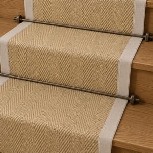 May include: A tan stair runner with a woven herringbone pattern and light grey fabric borders, secured to wooden stairs by two horizontal dark bronze metal rods with decorative end caps that span the width of the carpeted runner.