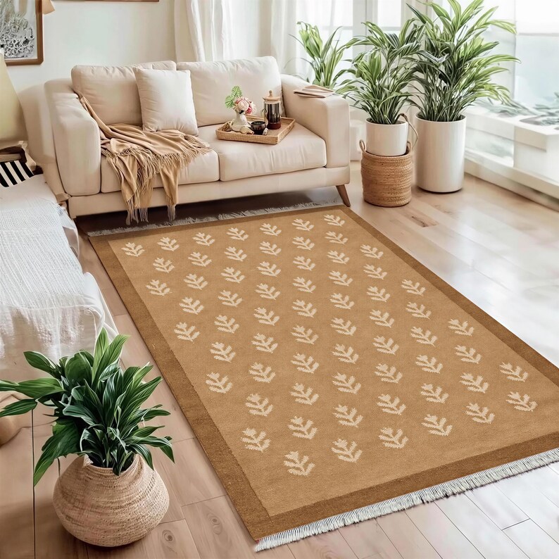 a cozy living room with a beige sofa, a large rug with a leaf pattern, and several potted plants. The room has a warm and inviting atmosphere, with a large window providing natural light.