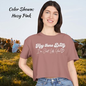 Puede incluir: Top corto rosa brumoso con el texto "Hey there Dolly, I'm Just Like You" en escritura blanca. La camiseta es de color liso y tiene cuello redondo. El color se describe como "Hazy Pink".