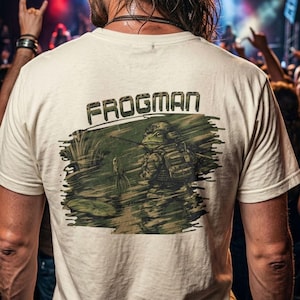 May include: A cream-colored t-shirt featuring a graphic on the back. The design shows a frog dressed in military gear, including a helmet and backpack, standing in a marshy pond. The text "FROGMAN" is printed in a blocky, camouflage-style font above.