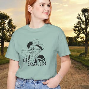 May include: A light blue t-shirt with a black and white illustration of a woman wearing a cowboy hat and holding a gun. The text "Gin Smoke & Lies" is printed below the illustration.