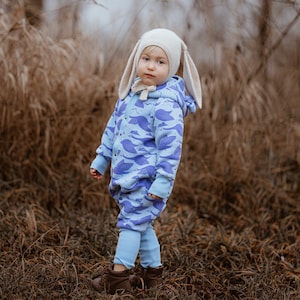 May include: A child in a light blue jumpsuit patterned with purple birds, light blue leggings, and brown boots. They wear a cream-coloured bunny hat with long ears. The background shows tall, dry grass. The outfit is suitable for a child.