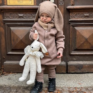 May include: A child wearing a brown bunny hat with long ears, a matching scarf, and a brown jumpsuit. The child is holding a white stuffed bunny with a ruffled collar. The child is wearing black boots.