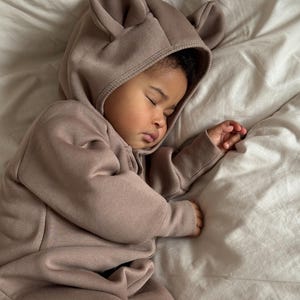 May include: A sleeping baby wearing a taupe-colored hooded jumpsuit with bear ears. The baby is lying on a white sheet. The jumpsuit has a zipper closure. The baby's eyes are closed, and the baby is resting peacefully.