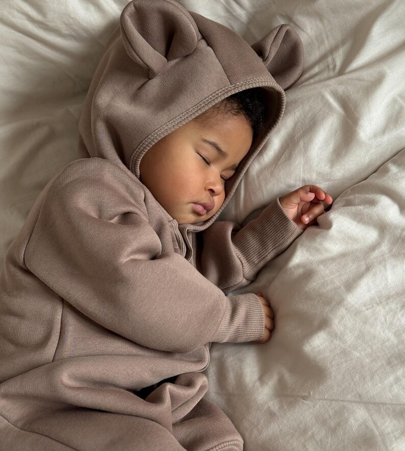 May include: A sleeping baby wearing a taupe-colored hooded jumpsuit with bear ears. The baby is lying on a white sheet. The jumpsuit has a zipper closure. The baby's eyes are closed, and the baby is resting peacefully.
