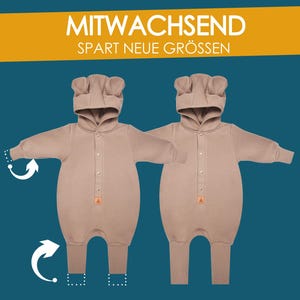 May include: Two tan-colored baby onesies with bear-shaped hoods. Each onesie has a button-up front and a small orange triangle label. The image includes the text "MITWACHSEND" and "SPART NEUE GR&Ouml;SSEN" in white letters on a yellow banner.