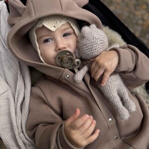 May include: A baby wearing a brown hooded jumpsuit with bear ears and a knitted bonnet. The baby is holding a knitted doll and a pacifier. The jumpsuit has snap closures and a small leather patch. The background is blurred.