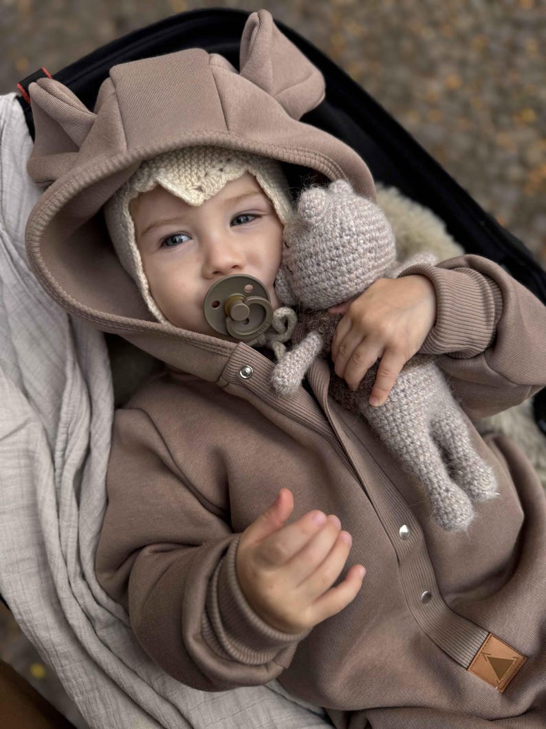 May include: A baby wearing a brown hooded jumpsuit with bear ears and a knitted bonnet. The baby is holding a knitted doll and a pacifier. The jumpsuit has snap closures and a small leather patch. The background is blurred.