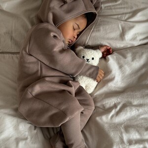 May include: A sleeping child wearing a brown hooded jumpsuit with bear ears, clutching a white teddy bear. The jumpsuit has ribbed cuffs and a front zipper. The child is lying on a bed with white sheets.
