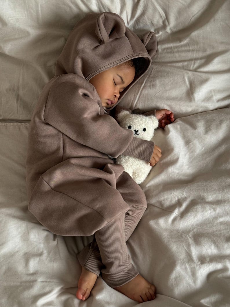 May include: A sleeping child wearing a brown hooded jumpsuit with bear ears, clutching a white teddy bear. The jumpsuit has ribbed cuffs and a front zipper. The child is lying on a bed with white sheets.