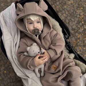 May include: A baby in a brown hooded jumpsuit with bear ears and a knitted hat, lying in a stroller. The baby holds a small gray crocheted teddy bear. The stroller is black, and the background is blurred.