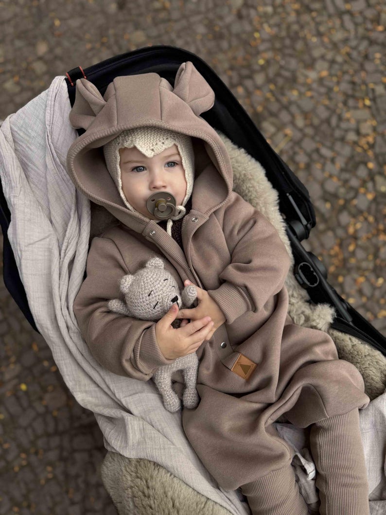 May include: A baby in a brown hooded jumpsuit with bear ears and a knitted hat, lying in a stroller. The baby holds a small gray crocheted teddy bear. The stroller is black, and the background is blurred.