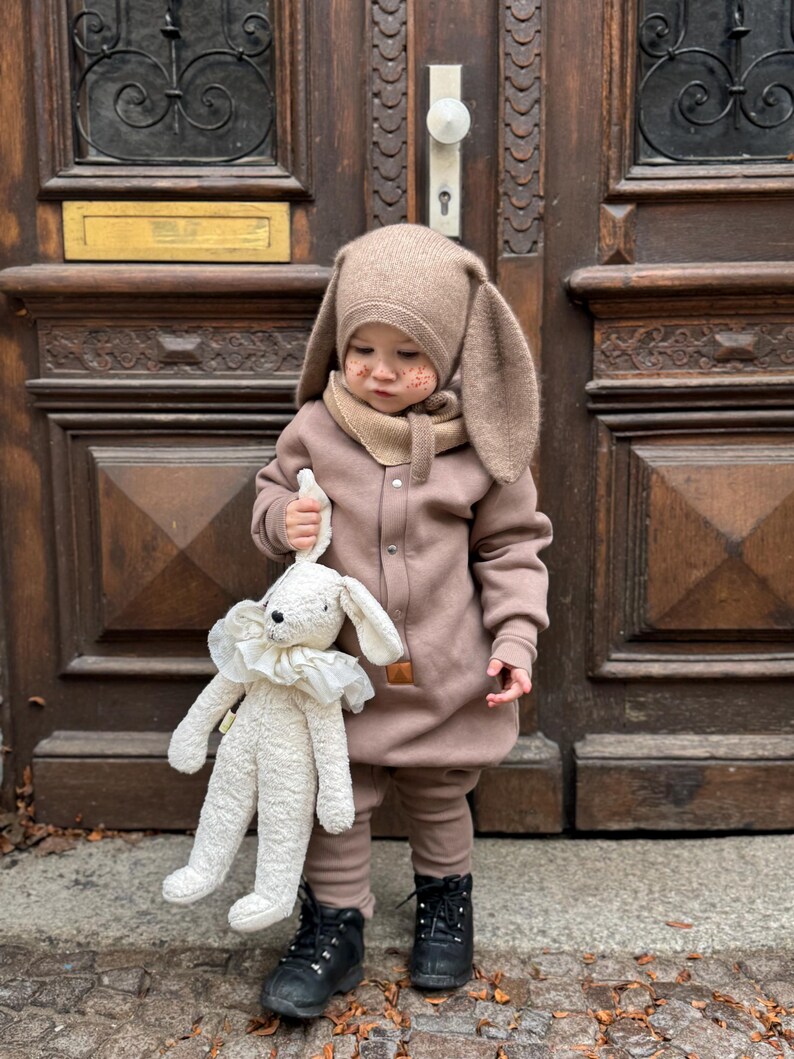 May include: A child wearing a brown bunny hat with long ears, a matching scarf, and a brown jumpsuit. The child is holding a white stuffed bunny with a ruffled collar. The child is wearing black boots.