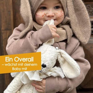 May include: A child wearing a tan bunny ear hat and a matching brown jacket, holding a white stuffed bunny. The image includes text in German that reads "Ein Overall - w&auml;chst mit deinem Baby mit."