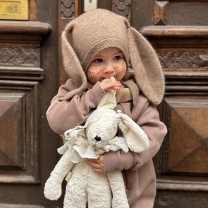 A child dressed in a hazelnut jumpsuit and a hat with ears stands in front of a door. He holds a stuffed animal in his hands.