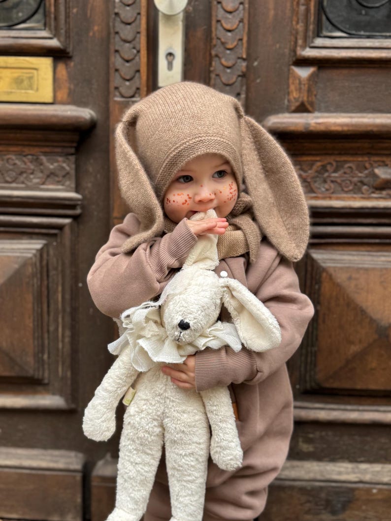 A child dressed in a hazelnut jumpsuit and a hat with ears stands in front of a door. He holds a stuffed animal in his hands.