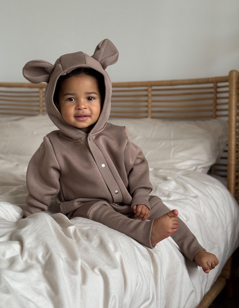 May include: A taupe-colored baby jumpsuit with a hood featuring bear ears. The one-piece outfit has a button-up front and ribbed cuffs on the sleeves and legs. The child is sitting on a white bed.