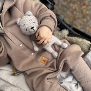 May include: A light gray crocheted teddy bear is held by a child wearing a tan hooded jumpsuit. The bear has a black nose and closed eyes. The jumpsuit has a ribbed cuff and a small leather patch.