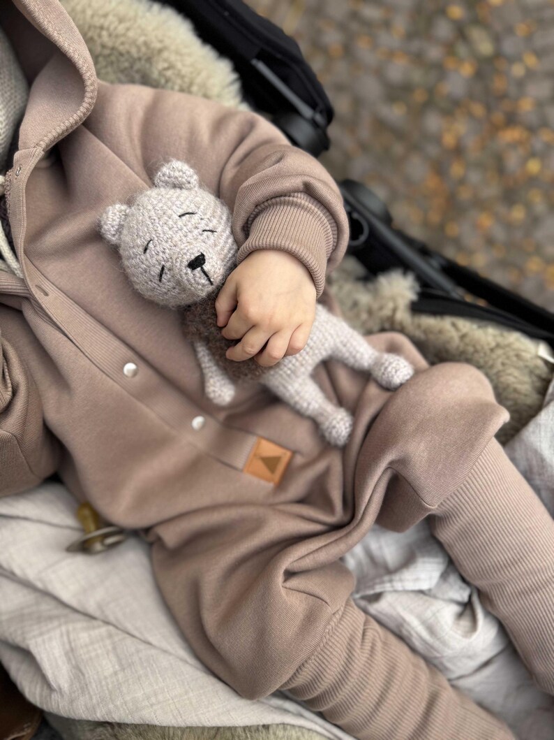 May include: A light gray crocheted teddy bear is held by a child wearing a tan hooded jumpsuit. The bear has a black nose and closed eyes. The jumpsuit has a ribbed cuff and a small leather patch.