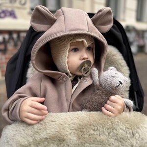May include: A baby wearing a tan hooded jacket with bear ears and a knitted hat, sitting in a stroller. The baby is holding a small, gray crocheted teddy bear and has a pacifier. The stroller has a faux fur lining.