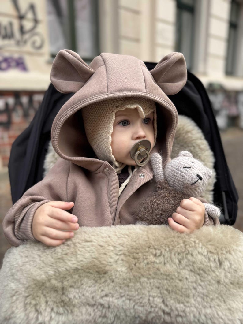 May include: A baby wearing a tan hooded jacket with bear ears and a knitted hat, sitting in a stroller. The baby is holding a small, gray crocheted teddy bear and has a pacifier. The stroller has a faux fur lining.