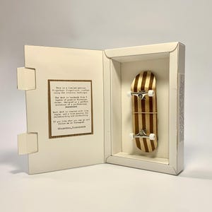 May include: A miniature skateboard with a striped wood design, presented in an open, off-white box. The skateboard has white wheels and silver axles. The box contains text that reads "This is a limited edition Fingerboard Fingerboard..."