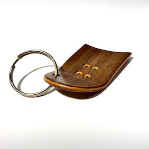 May include: A wooden fingerboard keychain with a silver key ring. The fingerboard is brown with a light brown wood grain and has five holes in the top.