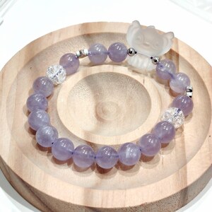 K&ouml;nnte beinhalten: Ein Armband mit runden, lavendelfarbenen Edelsteinen und klaren, facettierten Perlen. Es enth&auml;lt einen Charm aus mattiertem Glas in Form eines kleinen Tieres. Das Armband wird auf einer Holzoberfl&auml;che pr&auml;sentiert.