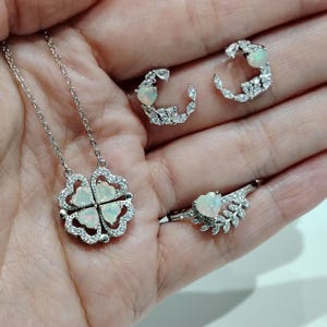 May include: A silver necklace, ring, and earrings set. The necklace features a four-leaf clover pendant with opal-like heart-shaped stones and clear crystals. The ring and earrings also have opal-like heart-shaped stones.