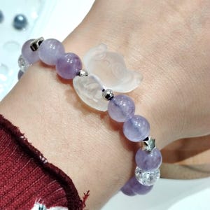 K&ouml;nnte beinhalten: Ein Armband mit lavendelfarbenen Amethystperlen, klaren Quarzperlen und silbernen Details. Es verf&uuml;gt &uuml;ber einen klaren, geschnitzten Schmetterlingsanh&auml;nger und einen silbernen Sternanh&auml;nger. Das Armband wird am Handgelenk getragen.
