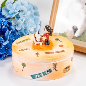 以下が含まれることがあります： 女の子と猫が描かれたカラフルなデザインのセラミック製の小物入れ。丸い形をしていて、蓋には立体的なデザインが施されています。猫が椅子に座っている、猫がフードボウルを持っている、猫が毛糸玉を持っているなど、様々な猫の画像で飾られています。